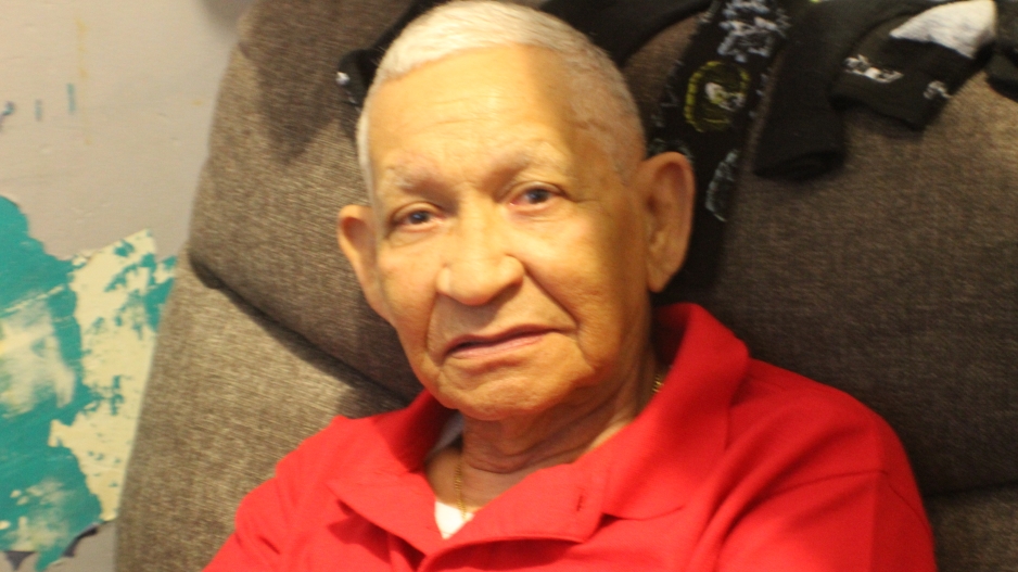 Image captures an older man with a head full of gray hair, sitting in his recliner. He looks directly, if not hesitantly in the lens of the camera.