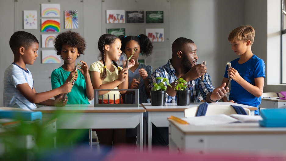 This image captures a teacher in a classroom working with five students. The teacher looks to engage five elementary school aged children. The surround the teacher seated at his desk. Each of the students are enthralled with their respective STEM assignments. Two of the the boys are paired together, while the two girls are also paired off with each other, letting their scientific curiosity get the better of them. One child is asks for help directly from the teacher.
