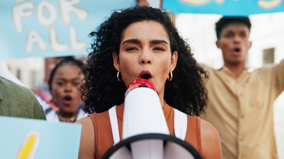 Image captures group protesting for improved education opportunities. The photo captures four protestors in the street bringing attention to their issue. The image features those holding cardboard signs and making noise to be disruptive. The photo focues on a latina woman holding a white megaphone as she chants in protest. She sports black should legnth hair, and wears a white top with vertical red strips.
