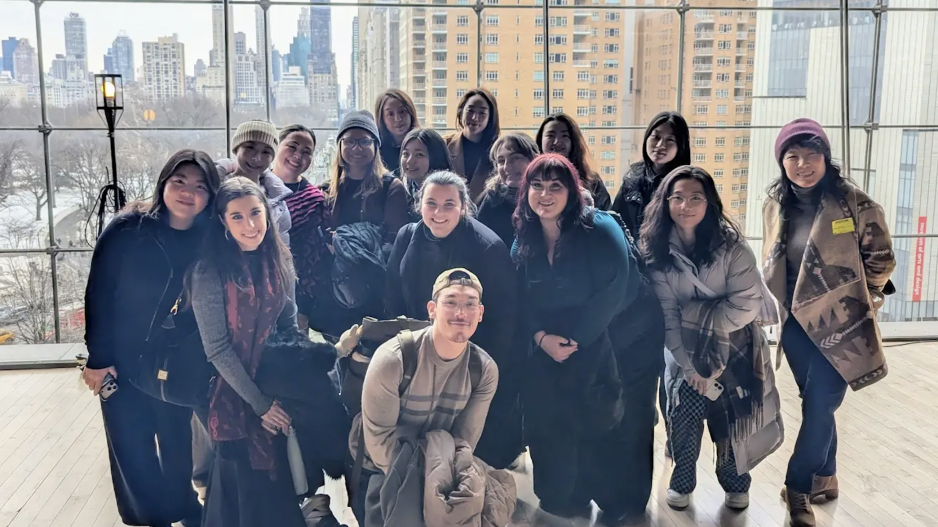 Group photo of NYU PAA students inside the Appel Room with the Manhattan skyline visible through the large windows behind them.