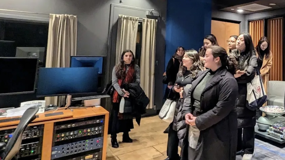 Students listen to a staff member speaking in a recording studio, with equipment and monitors visible nearby.