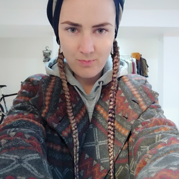 me dressed in a coat with geometric patterns, with two long braids sticking out of the scarf wrapped around my head. I'm looking right at the viewer, slightly and cunningly smiling 