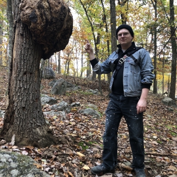 Disheveled man in a jean jacket points at a tree burl