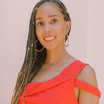 Photo of Heather Clarke a Brown Skinned Black monoracial Autistic woman with long waist length braids wearing a read top and gold hoop earrings. She is looking ahead at photo and smiling. 