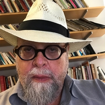Allen wears black rimmed glasses and a fedora hat. He has a white beard and stands in front of his office bookcase. 