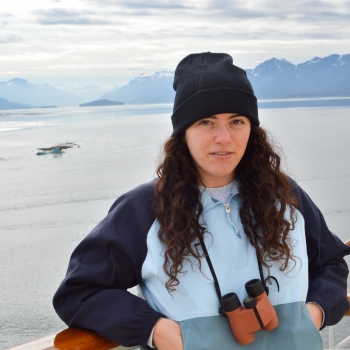 Profile picture. Woman with long, brown curly hair wearing a black beanie. Standing in front of a background of mountains and water.