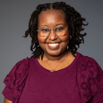 Headshot of Britton Williams: smiling Black woman with black locs against a gray backdrop. She is wearing gold glasses and a burgundy shirt.