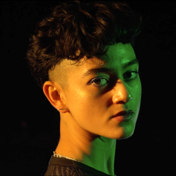 Image of Luna Beller-Tadiar, a queer person with short black hair, shaved sides, and light brown skin. She is lit by green and orange light and looks at the camera.