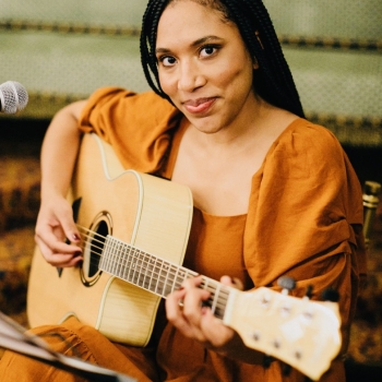 Image of a Black woman with box braids looking at the camera and smiling while holding a guitar, wearing an orange dress