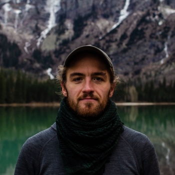 A photograph of Sam Kellogg, a light-complexioned man looking into the camera. He is wearing a green baseball cap with a bird on it, a green scarf, and a grey shirt, standing in front a blue-green mountain lake, a dark green evergreen forest, and grey cliffs.