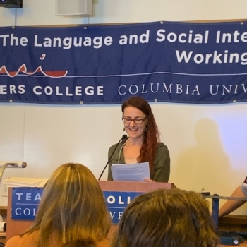 Sasha stands at the podium at a conference on Language and Social Interaction at Teacher's College, Columbia. She has red slightly wavy hair sliding down on one side of her head, she is smiling and reading a presentation from her notes