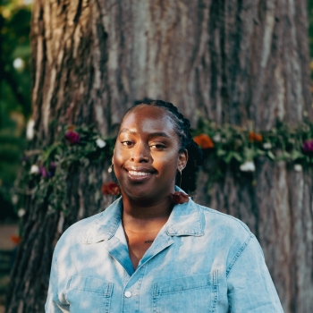 A black skinned woman in a denim jumper smiles at the camera. Her hair is pulled back. She is outside standing in front of a large tree trunk.