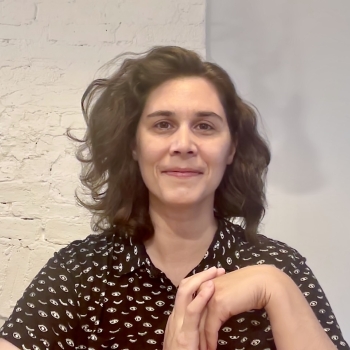 Image is of a white person with shoulder length wavy hair, parted to one side, wearing a black collared shirt with a repetitive white pattern.