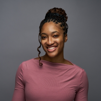 Olivia O'Leary with braided hair in a bun, wearing a mauve long-sleeve top, smiles in front of a plain gray background.