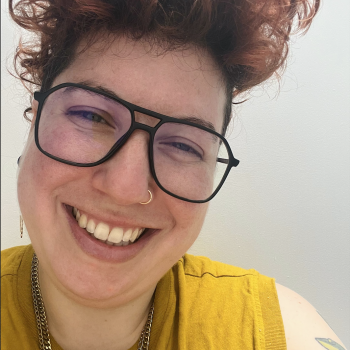 In this image, Roxy has short red curly hair, black aviator glasses, and a yellow shirt.