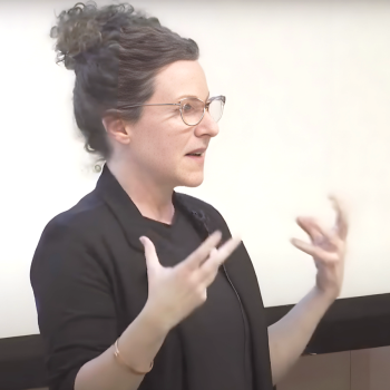 A white woman in a black blazer holds her hands in mid air at the podium. In the corner left screen is a woman interpreting using sign language. 
