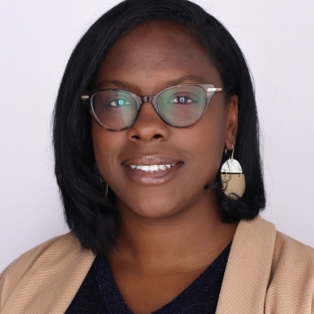 A black woman in glasses and a tan blazer smiles at the camera