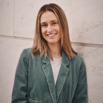A woman in a teal blazer with shoulder length blond hair leans against a wall and smiles at the camera
