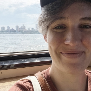 A white woman in her 30s with short hair, in front of a window with a skyline view.