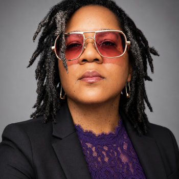 African-American woman with rose tinted glasses and locs wearing a black blazer and violet lace top