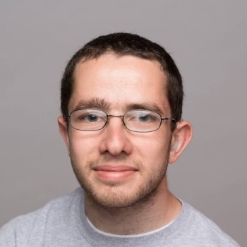 Visual Description  Subject: A young man with short brown hair, wearing metal-rimmed eyeglasses and a light gray crewneck shirt.  Expression: A subtle, gentle smile with a neutral gaze directed forward.  Background: A solid, plain light gray studio backdrop.