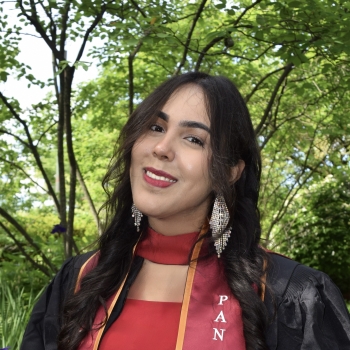 A young woman is shown in front of some trees with long dark hair, large dangling earrings, and has a graduation sash around her neck.