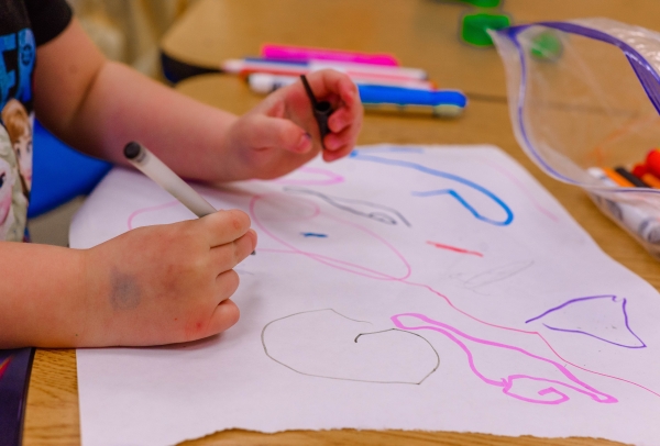 A photo of the hands of a child drawing scribbles with multicolored markers on a piece of paper
