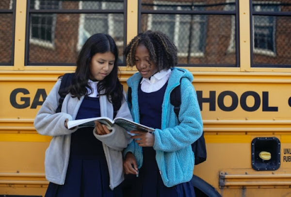 Two girls hold a textbook and look at the pages as they stand in front of a school bus