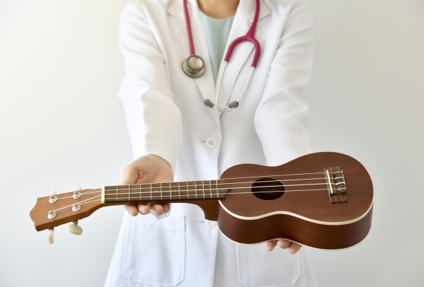 A doctor in a lab coat with a stethoscope around their neck holding out a ukulele with extended arms