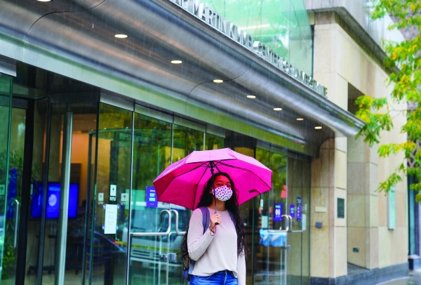 A student stands in front of the Kimmel Center under a pink umbrella. It is raining and she is wearing a face mask.