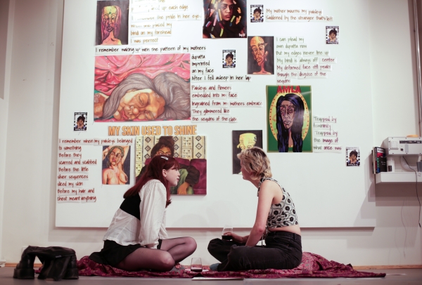 Two students sit on a blanket in front of a large canvas. The students are facing each other and sipping red wine out of stemless glasses.