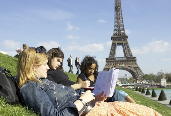 Three students are sitting on the lawn in front of the Eiffel Tower and reading a notebook
