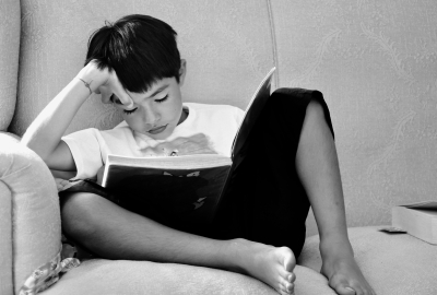 A child sitting on a couch and reading a book with his hand on his forehead