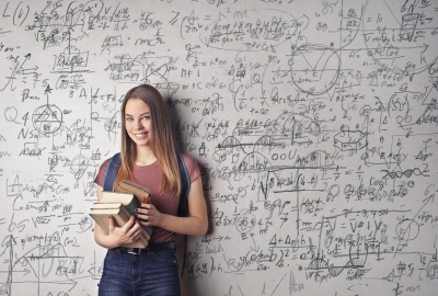 A young woman holding books standing in front of a dry erase board covered in math equations