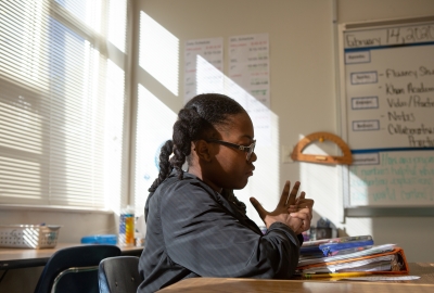 A Black girls sits alone in a classroom staring down at her assignment