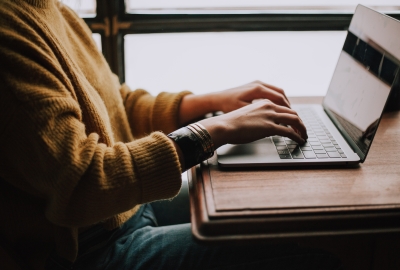A stock image of a person in a sweater typing on a laptop. Their face is cropped out of the photo.