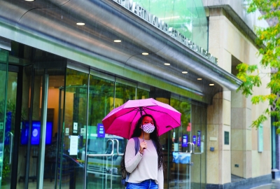 A student stands in front of the Kimmel Center under a pink umbrella. It is raining and she is wearing a face mask.