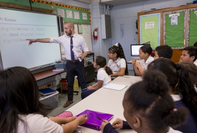 A teacher is standing at the front of a classroom filled with students in uniforms. He is pointing at the screen, which is showing a review of the content.
