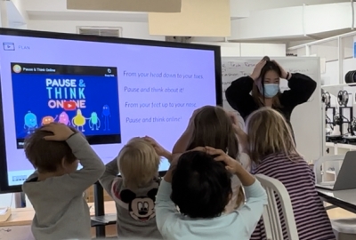 Teacher in face mask leading a classroom of young children in front of a TV projector displaying a slide with a video entitled "Pause & Think Online." The children and teacher have their hands on their heads in what seems to be a game or physical exercise. The children have their backs to the viewer. 