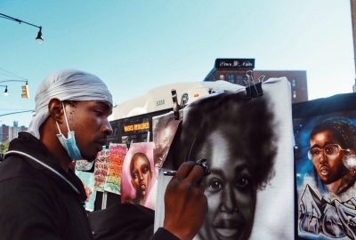 Street artist airbrushing a portrait of a Black woman.