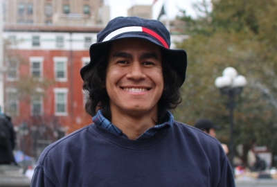 Photo of Ben in Washington Square Park; smiling, wearing a hat and navy blue sweater