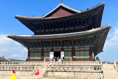 A view of Gyeongbokgung Palace in Seoul, with several people in brightly colored clothing in front of the palace.