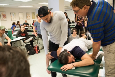 A man, laying facedown on a padded medical table, has his arm manipulated by a physical therapy student while an instructor and classmates look on.
