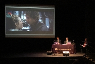 Screen showing Star Wars clip with seminar speakers sitting at desk in front of it discussing
