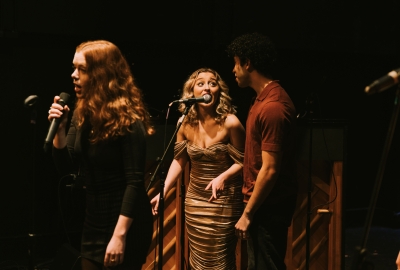 Three singers on stage performing with piano in background