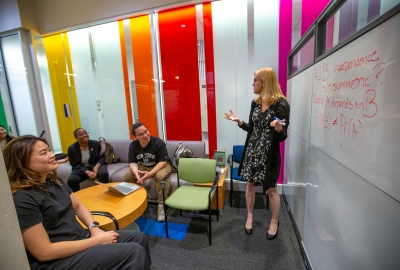 Professor Jennifer Hill by white board, explaining an equation to 3 sitting students 