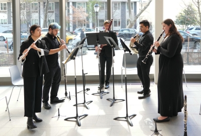 Five brass and woodwind performers playing in front of large sunny windows