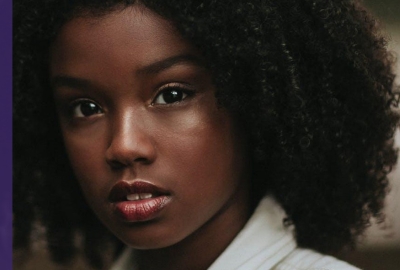 Image captures a Black girl with curly black hair looks pensively into the camera.