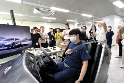 A man in a mask sits in a small, open car inside a lab, surrounded by other students