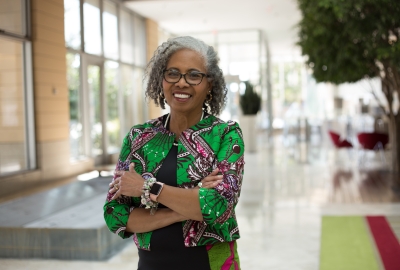 Image captures photo of Gloria Ladson-Billings.  The photo focuses on Dr. Ladson-Billings standing with arms folded against her person. She wears a green and pink colored top and eye-glassed. She is the only person featured in the image. 